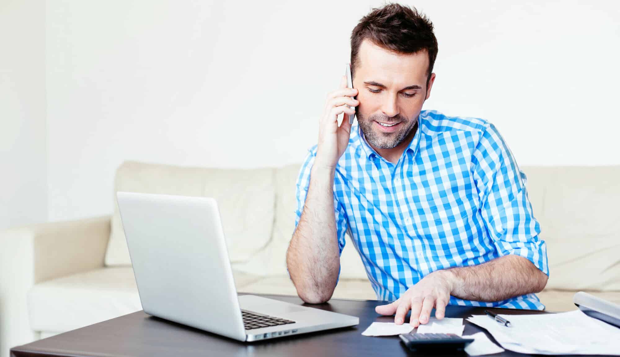 Man in blue plaid shirt working on a calculator and papers on laptop