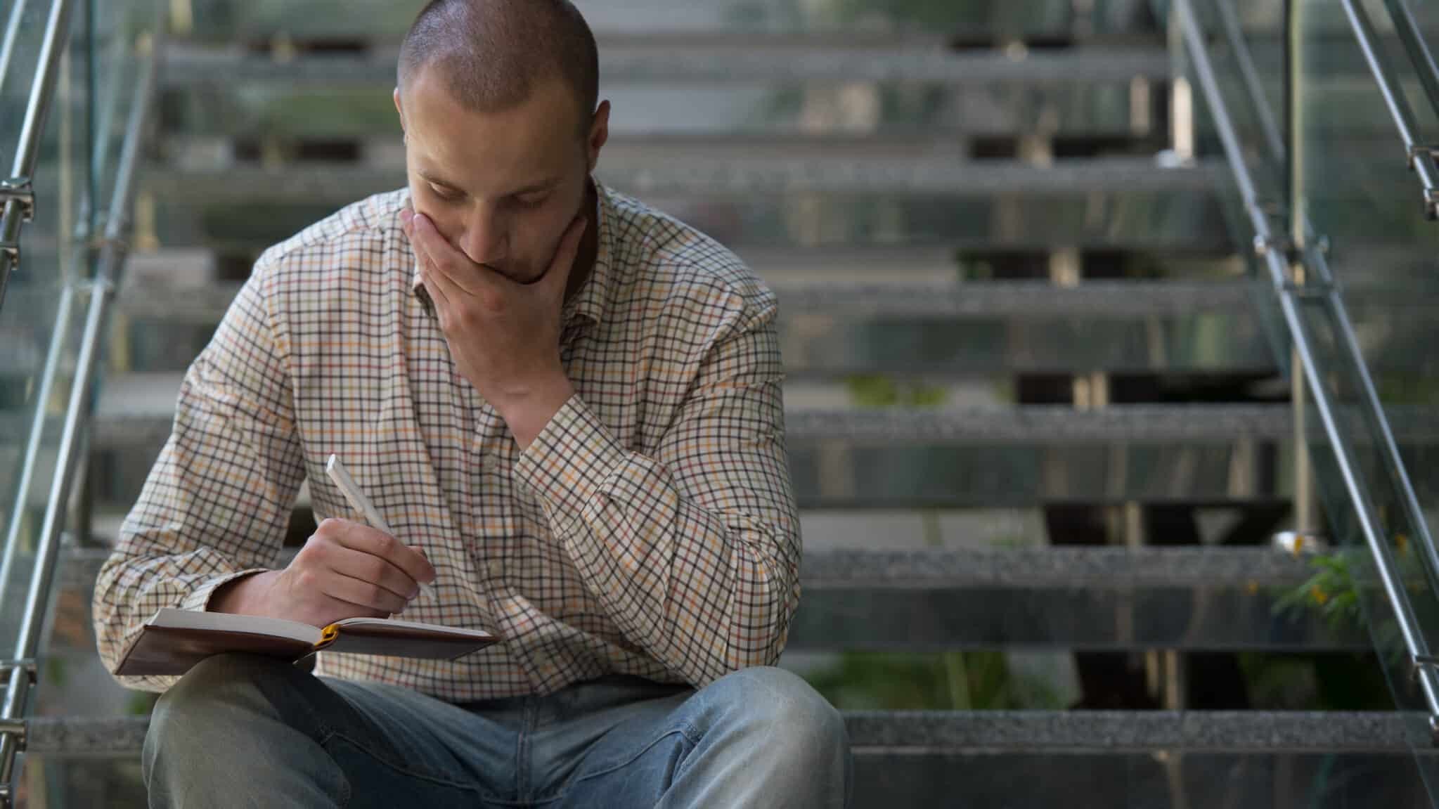 A man sitting on steps, writing on a pad of paper and looking determined.