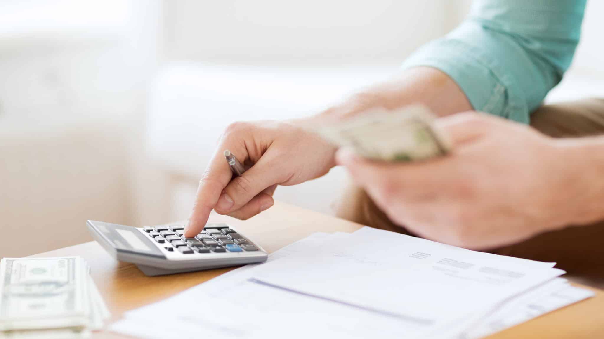 A person using a calculator to budget their finances, with papers and cash on the desk.