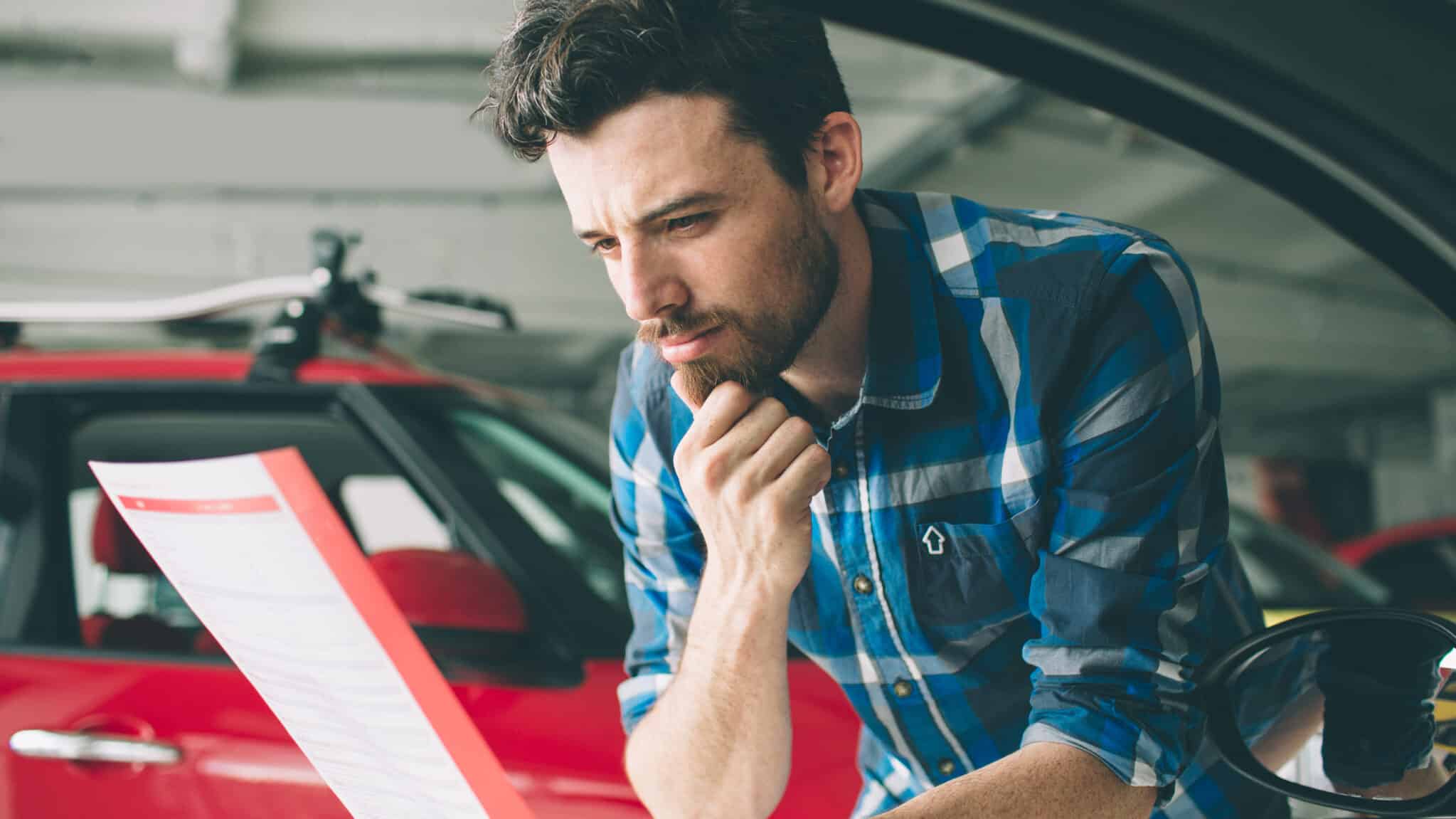 A young man sitting at a desk, looking at paperwork, with a new red vehicle parked in the background.