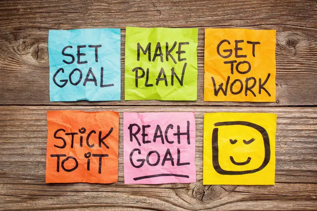 six colorful sticky notes on table that say set goal - make plan - get to work - stick to it - reach goal - smiley face 