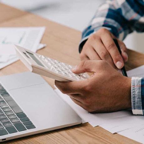 Person using a calculator at a desk with a laptop and papers, possibly managing finances or doing taxes.